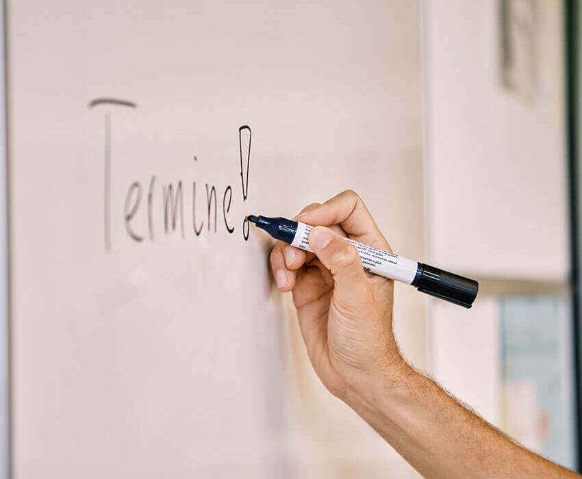 Eine Hand schreibt mit einem Stift etwas in einen Kalender und die zweite hält ein Handy