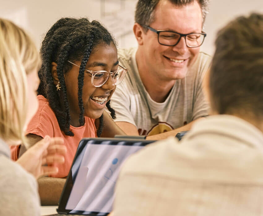 Ein Lehrer sitzt mit Schülern an einem Tisch und sie lachen gemeinsam währen sie am Tablet lernen