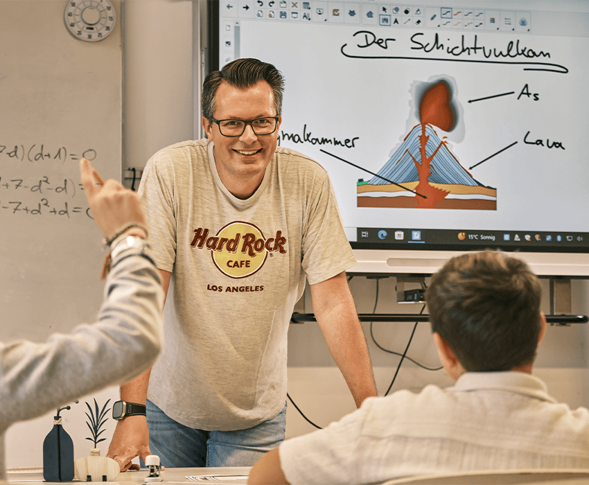 Ein Lehrer steht vor der Tafel und lächelt in die Kamera, die Schüler sitzen auf ihren Plätzen und melden sich
