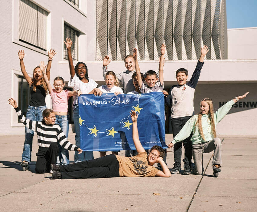 Eine Gruppe von Schülern steht vor der Schule und hält eine Erasmus Flagge in die Luft und ihre Hände
