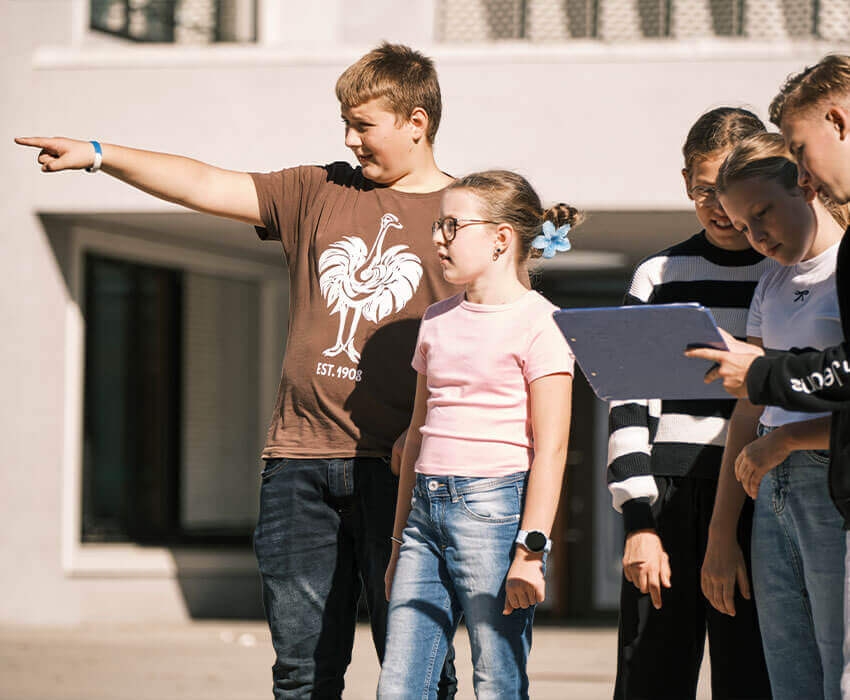 Ein Junge zeigt einem Mädchen etwas, hinter ihnen steht eine Gruppe an Schülern die sich etwas auf einem Clipboard ansehen.