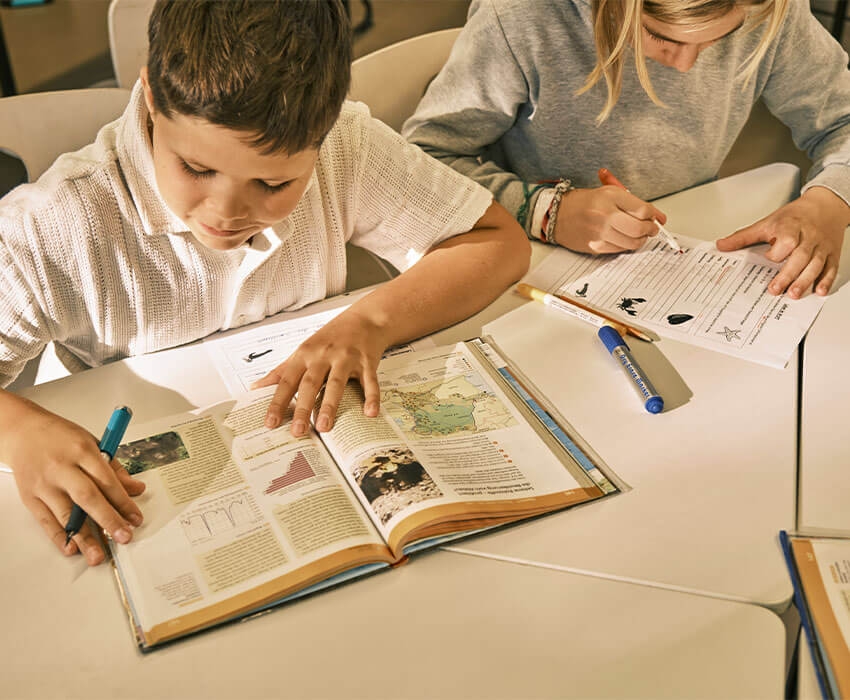 Zwei Kinder sitzen an ihren Schulheften und bearbeiten die Aufgaben darin.